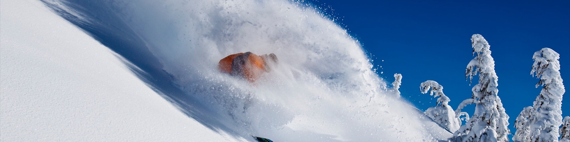 A snowboarder carving through deep snow with a clear blue sky in the background.