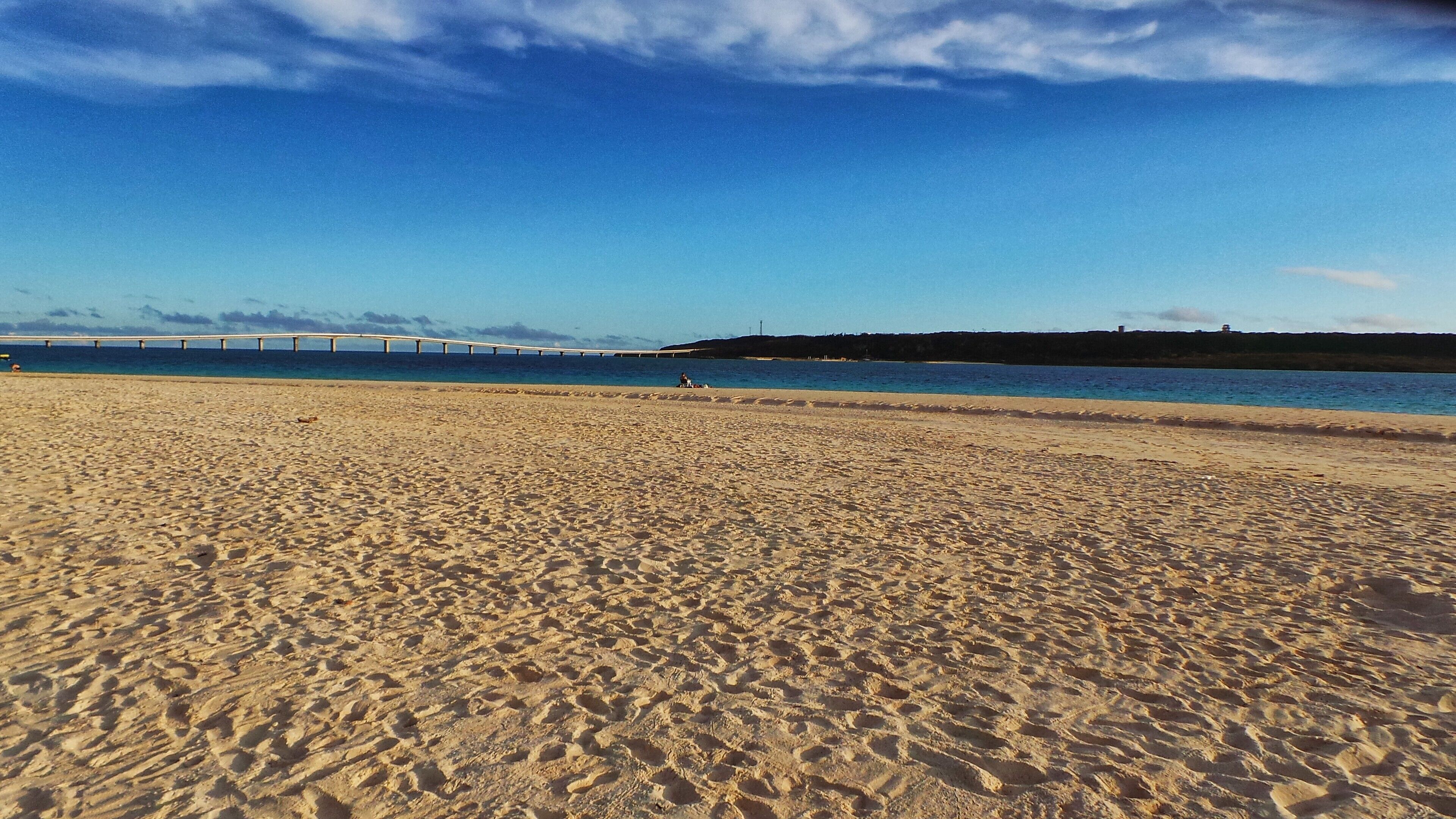 A sandy beach with footprints, a person sitting, and a bridge in the background.
