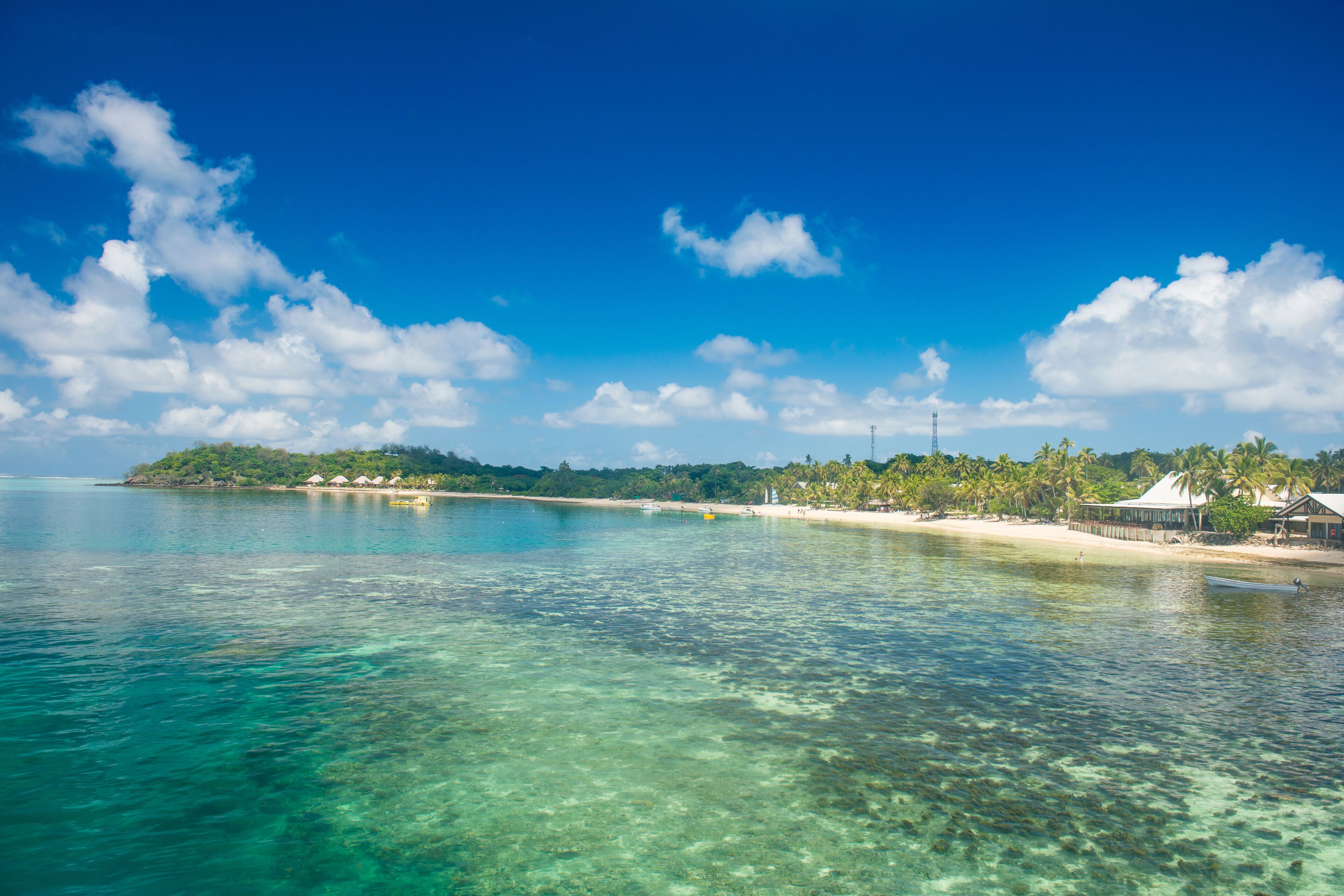 Una playa tropical con aguas turquesas cristalinas, una orilla de arena y una pequeña isla con vegetación y edificios.