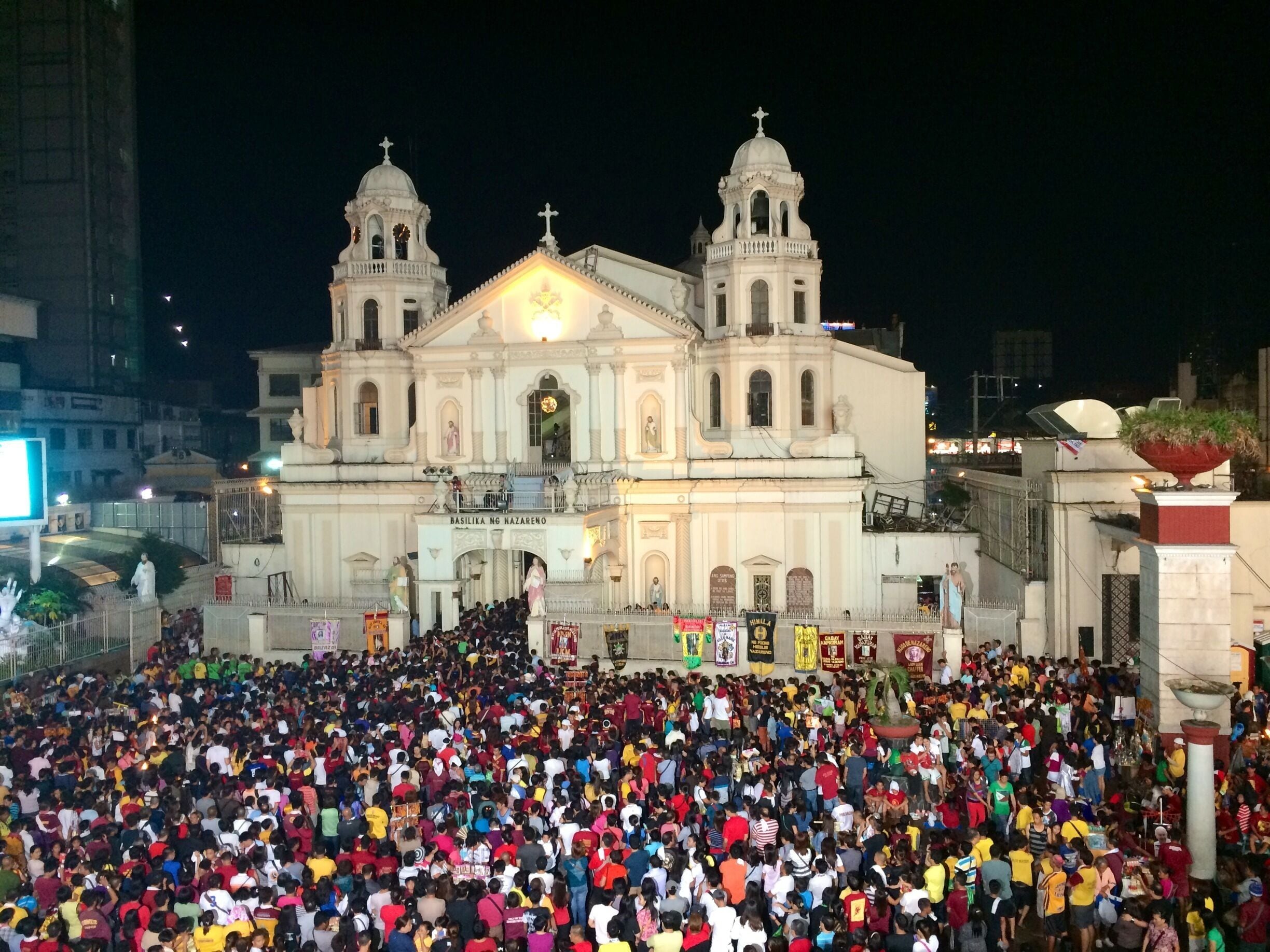 Uma grande multidão reunida em frente a uma catedral imponente com duas torres e uma cúpula central, sob um céu noturno.