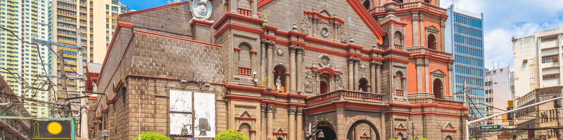 A historic church with red and grey architecture, a tuk-tuk on the street, and modern buildings in the background.