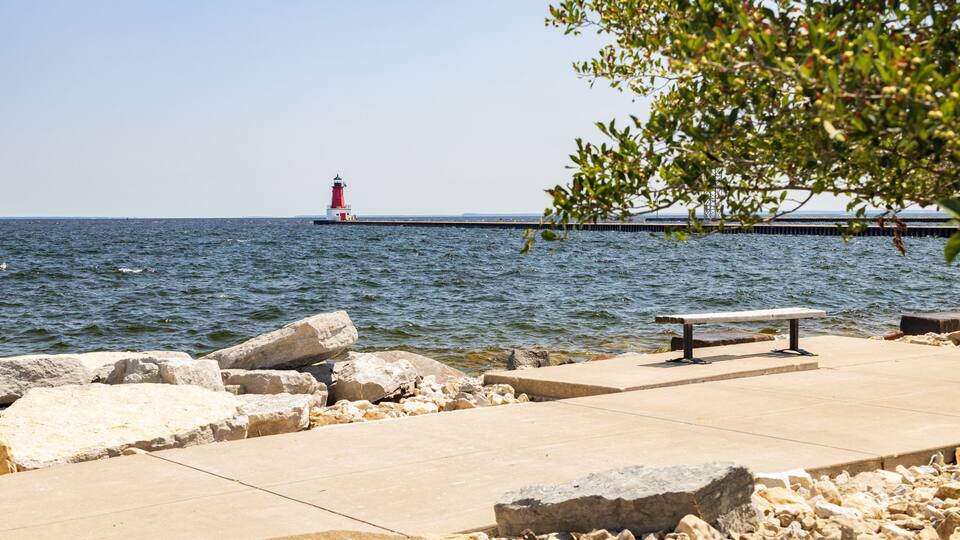 A lighthouse, rocky shoreline, and a bench on a concrete pier.