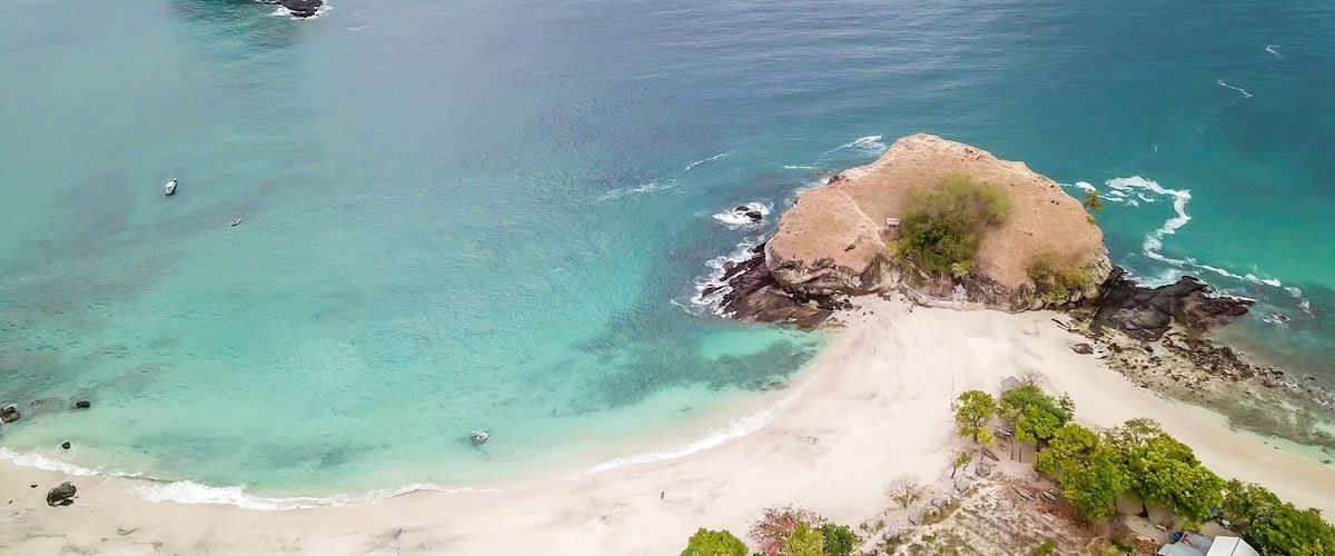 A coastal landscape with clear turquoise waters, a sandy beach, and a rocky outcrop with vegetation.