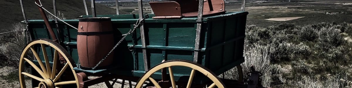 A green wagon with large wooden wheels and metal arches, set against a mountainous landscape with a clear sky.
