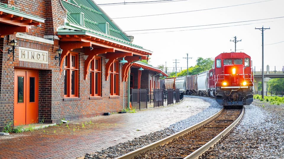 A red train approaching a brick building with a green roof and the sign 'MINOT.'