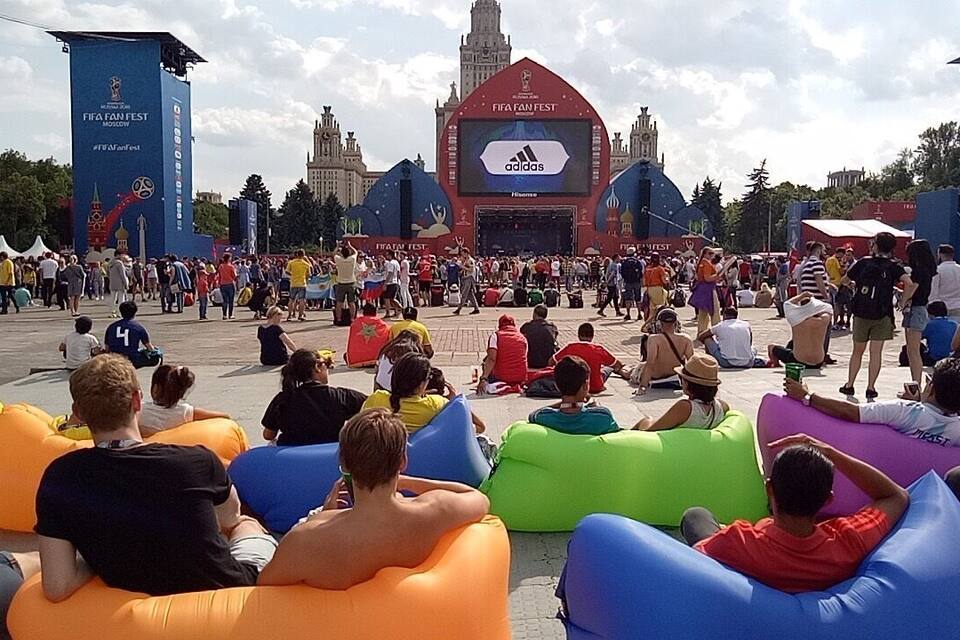 Uma praça pública movimentada com assentos infláveis e uma tela grande exibindo o logotipo do FIFA Fan Fest.