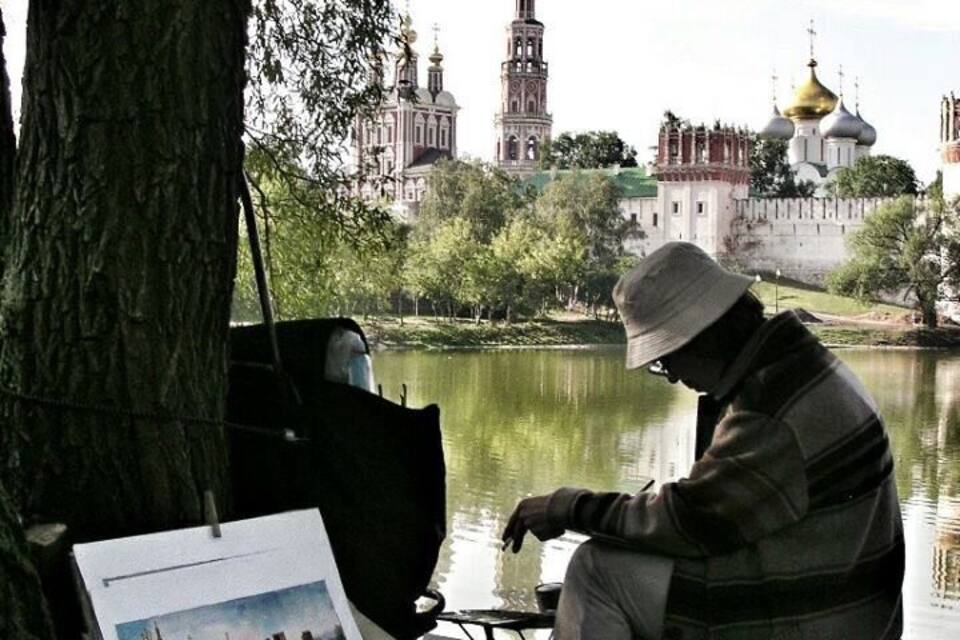 Uma pessoa sentada à beira de um lago, com um laptop e papéis, árvores e uma vista distante de um edifício histórico com cúpulas.