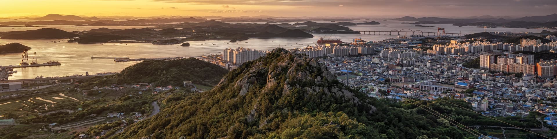 A coastal cityscape with a mountain in the foreground, a bridge in the distance, and a sunset sky.