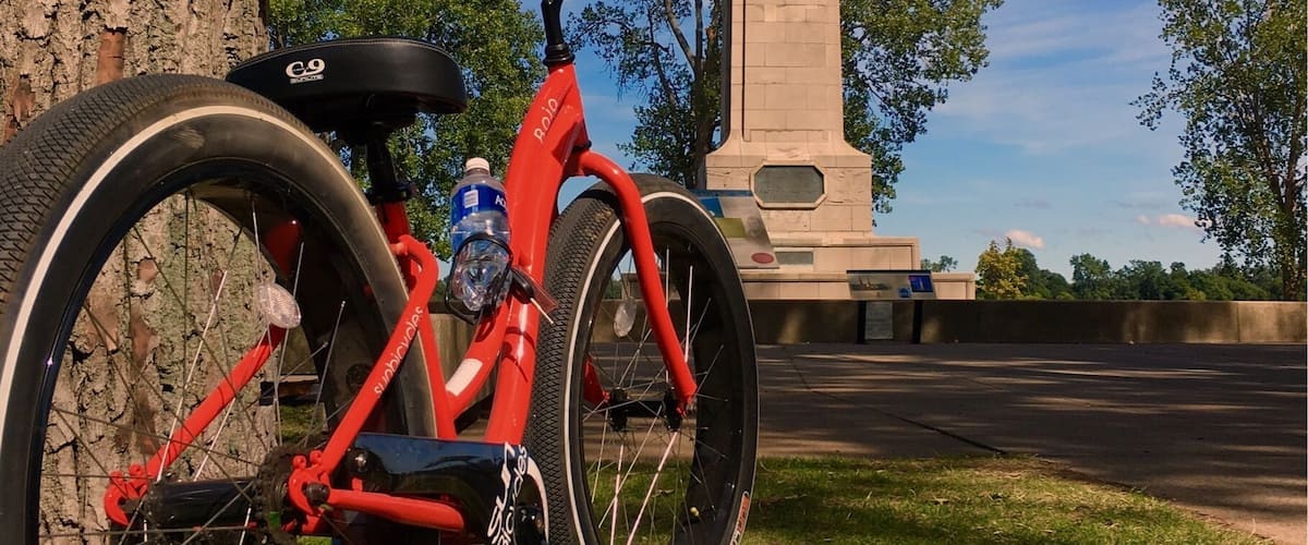 A red bicycle parked near a tree, with a tall monument in the background.