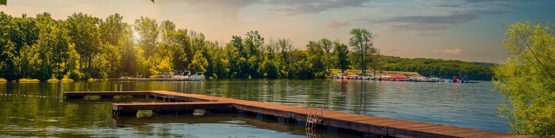 Un lago tranquilo con un muelle de madera que se adentra en el agua, rodeado de frondosos árboles verdes y barcos anclados a lo lejos.