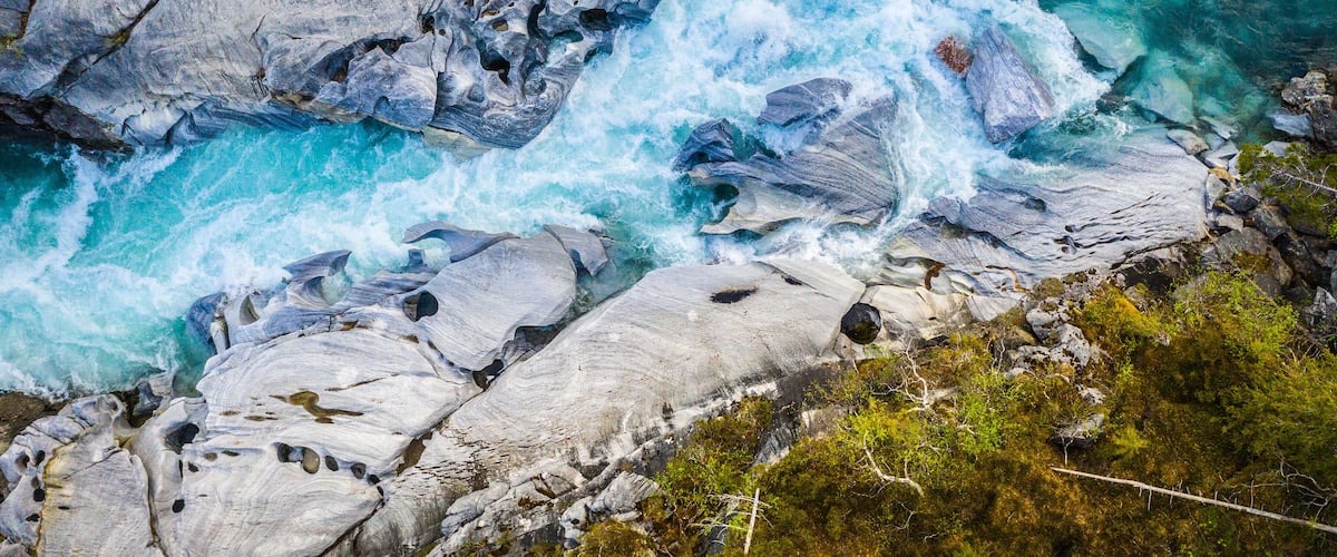 A turbulent river flowing over and around large rocks, surrounded by greenery.