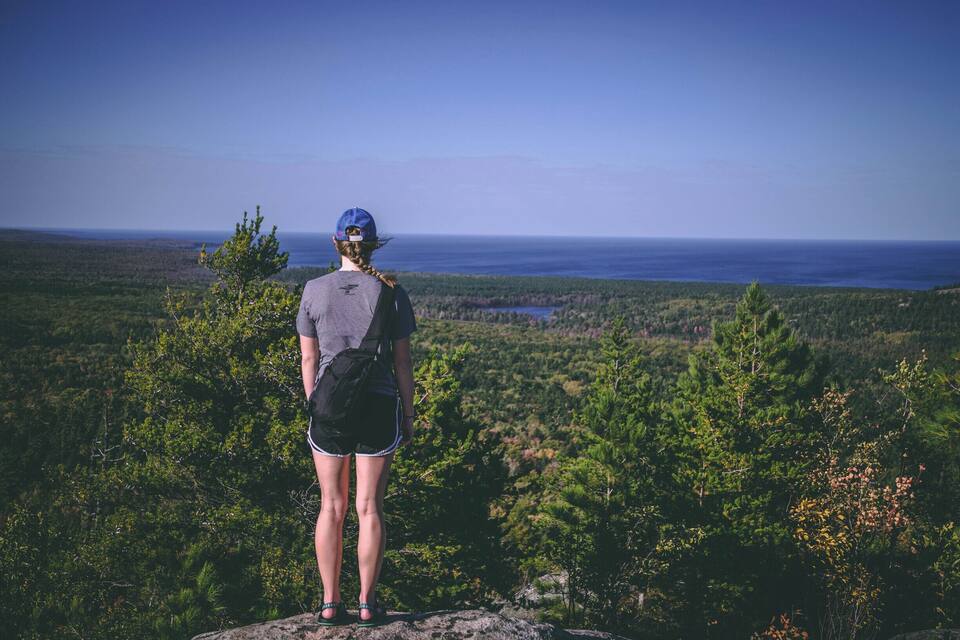 Una persona de pie sobre un afloramiento rocoso, con vistas a un bosque frondoso y una masa de agua.