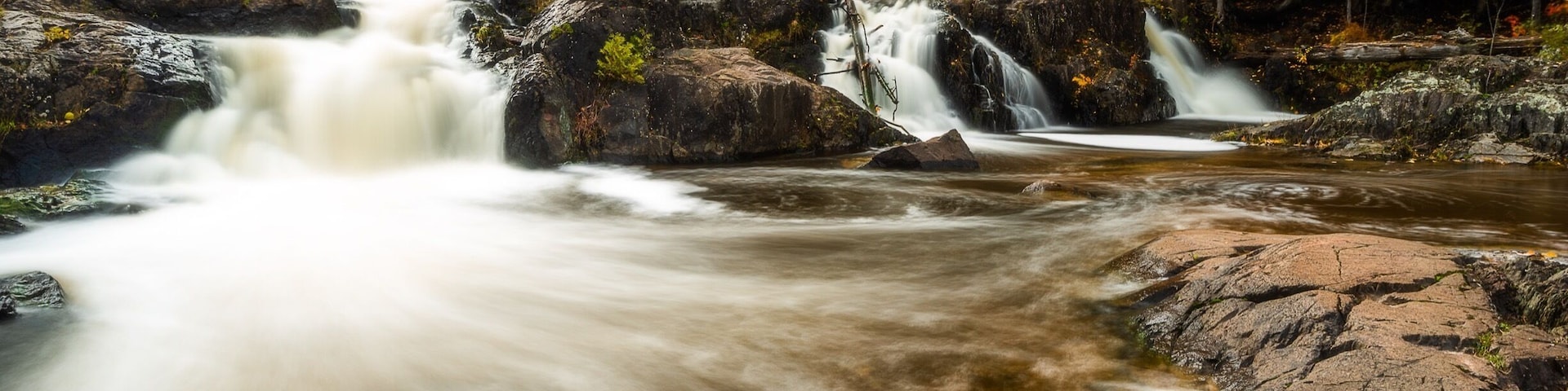 Una cascada se precipita sobre rocas en un área boscosa con árboles de colores otoñales.
