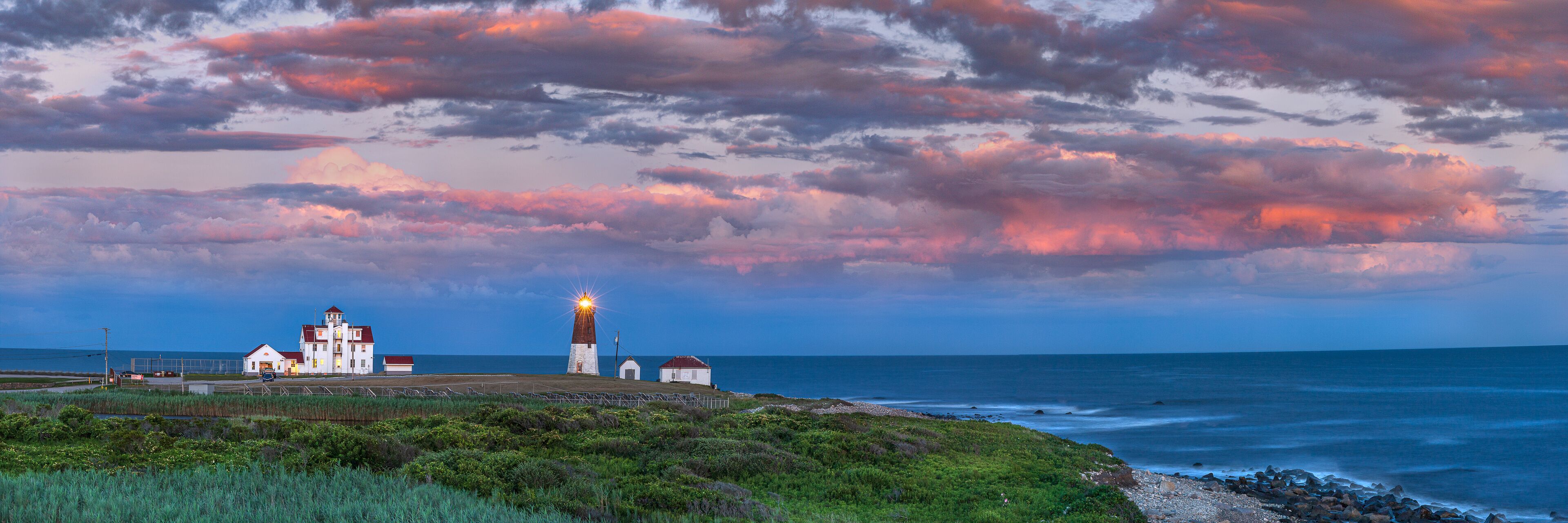 Un faro costero con techo rojo, rodeado de terreno cubierto de hierba y un litoral rocoso.