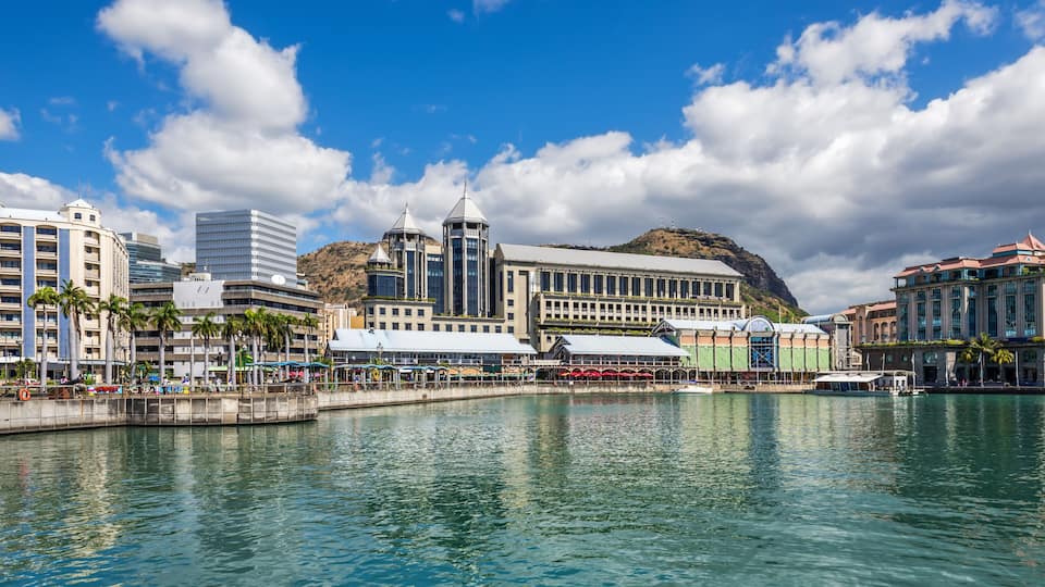 A waterfront cityscape with a prominent building featuring a tower, palm trees, and a hill in the background.