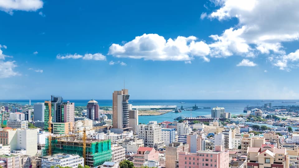 A coastal cityscape with a mix of modern and historic buildings, a prominent tower, and a view of the sea and a bridge in the distance.