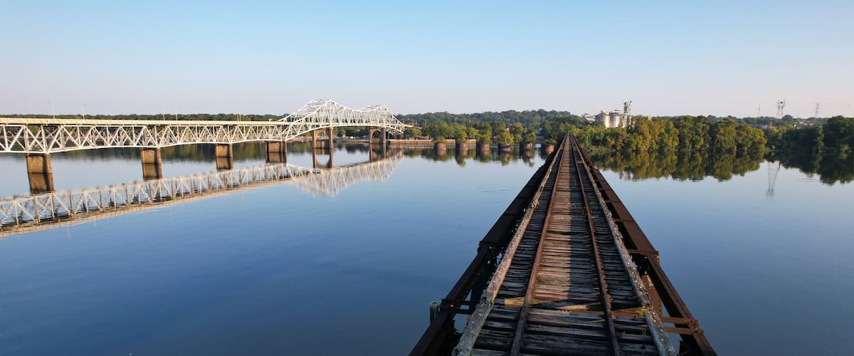 A railway bridge over calm water with a forested landscape in the background.
