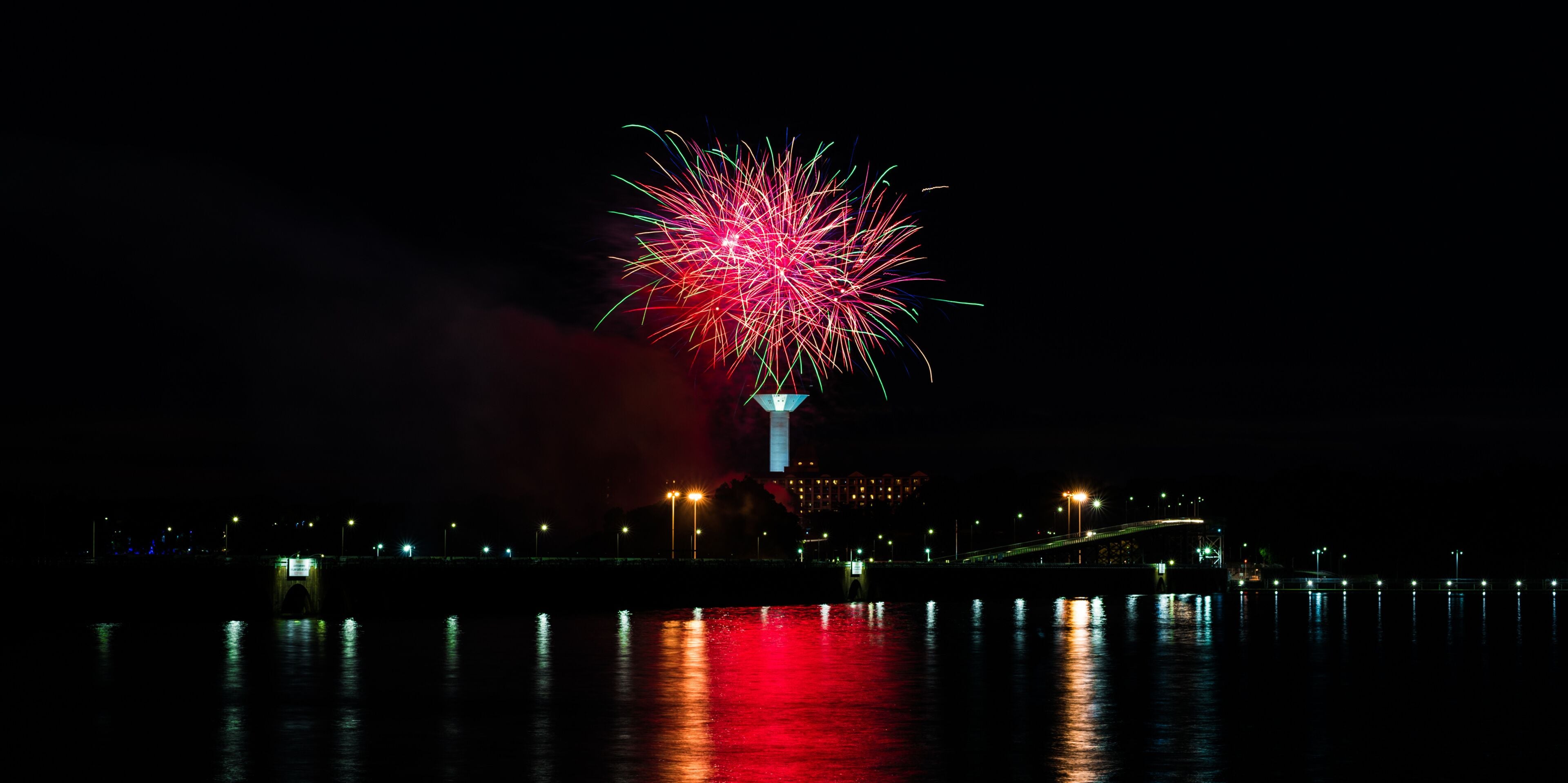 A night scene with a fireworks display over a body of water, a bridge, and a building with illuminated windows.