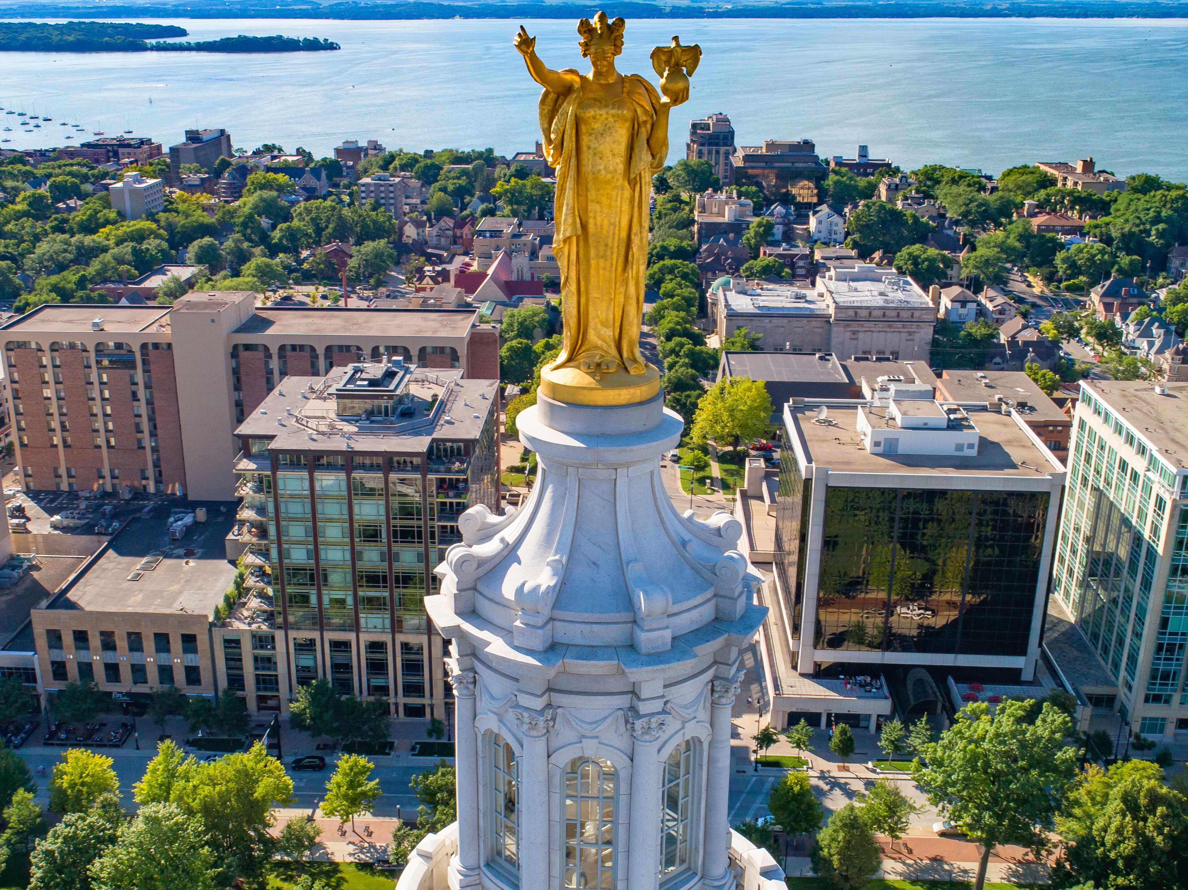 Una estatua dorada en lo alto de una columna blanca, con vistas a un paisaje urbano con edificios y una masa de agua.