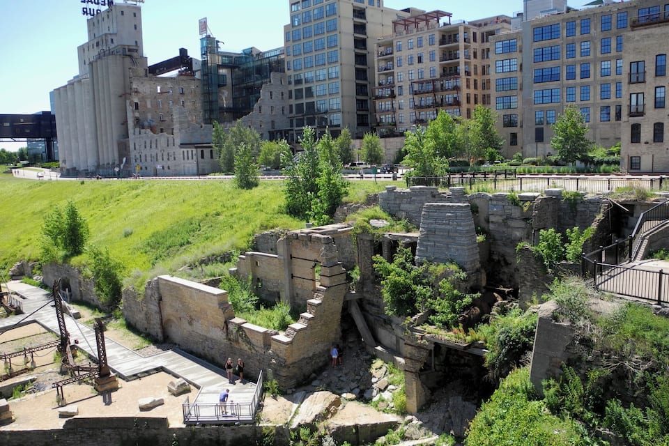 A historical site with ruins, a river, and modern buildings in the background.
