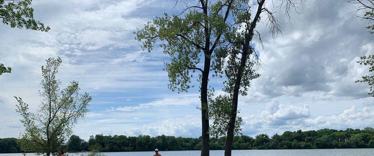 A lakeside beach with people relaxing, trees, and inflatable rafts.