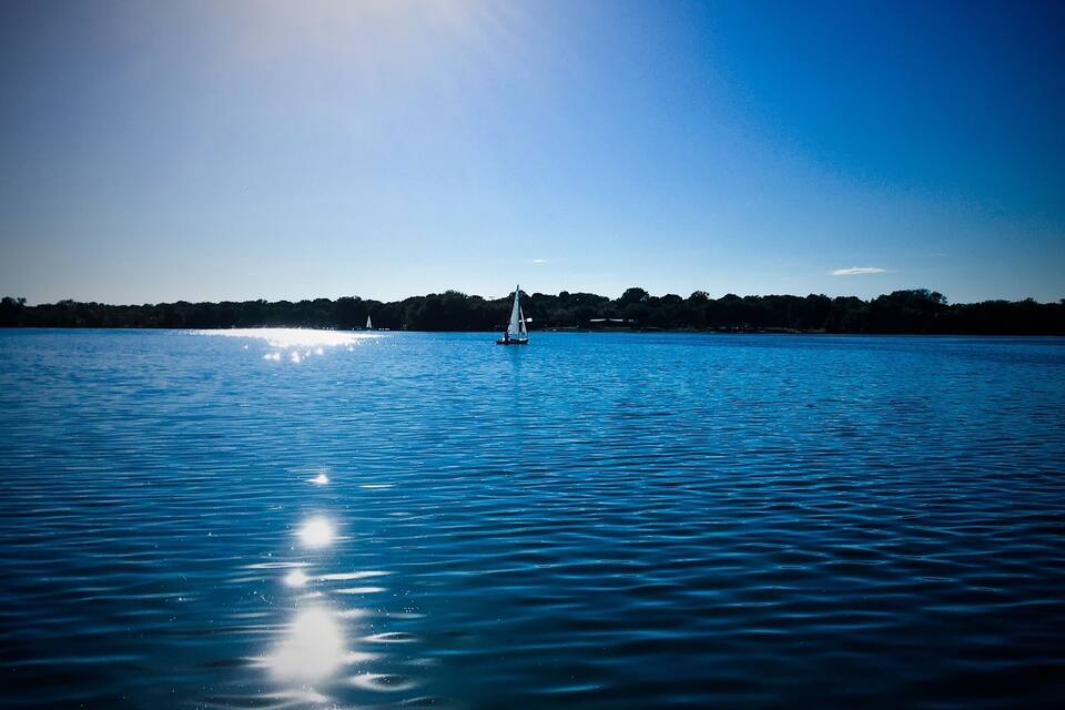 A calm lake with a sailboat in the distance, surrounded by a forested shoreline.