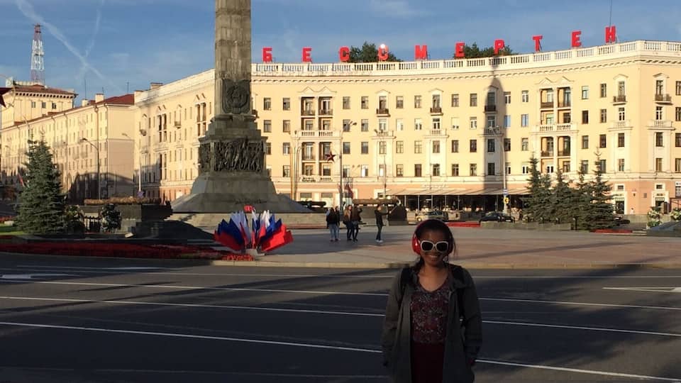 A person standing in a square with a monument and a building with 'EECM' on the facade.
