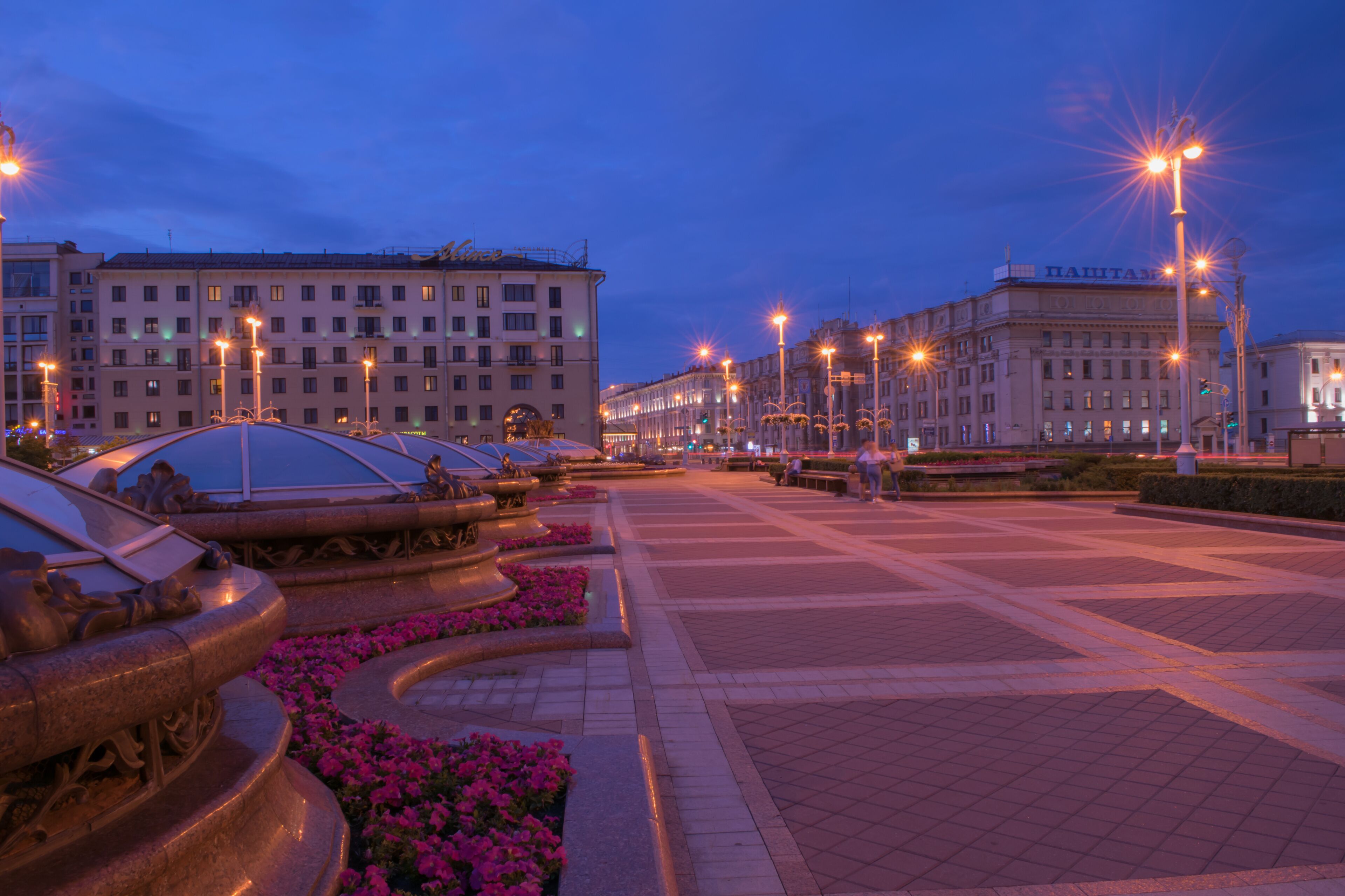 A city square with ornate fountains, flower beds, and classical architecture.