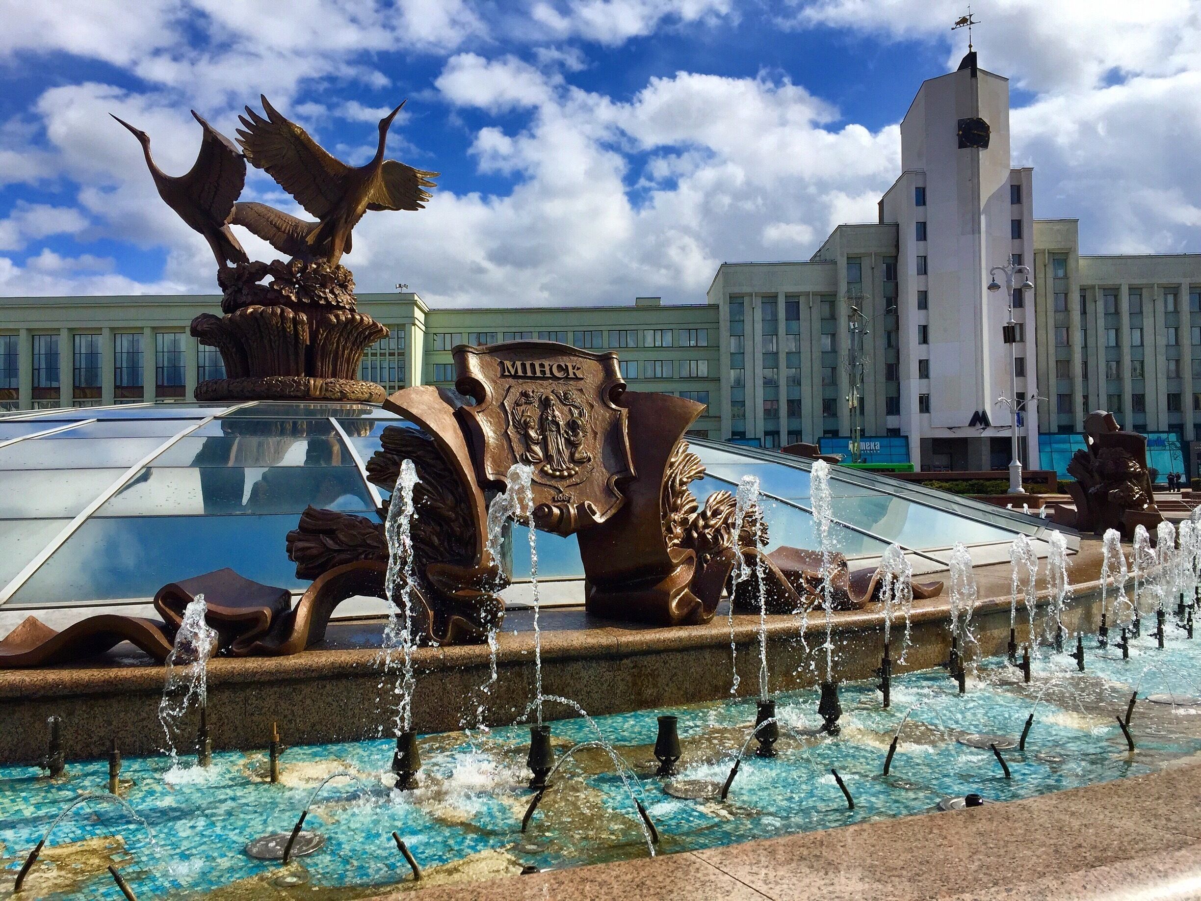A fountain with water jets and a statue of a bird. In the background, there is a building with the word "MIFLICK" on it.