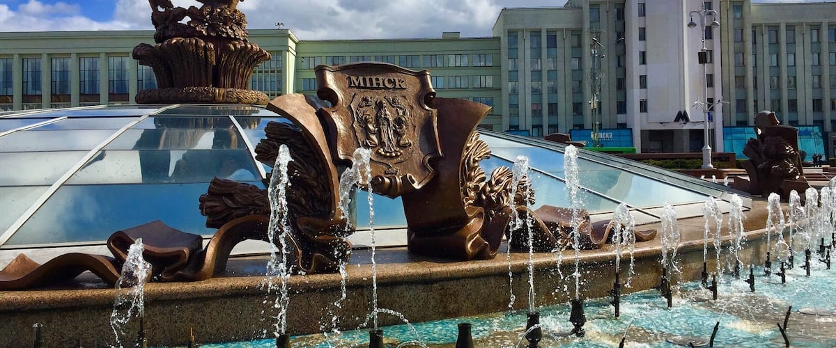 A fountain with water jets and a statue of a bird. In the background, there is a building with the word "MIFLICK" on it.