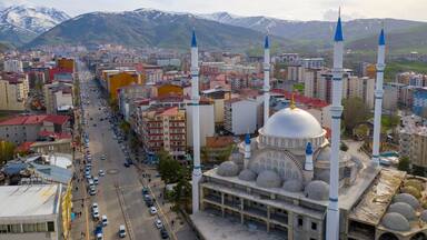 A cityscape with a prominent mosque featuring a large dome and two minarets, surrounded by urban buildings and a mountainous backdrop.