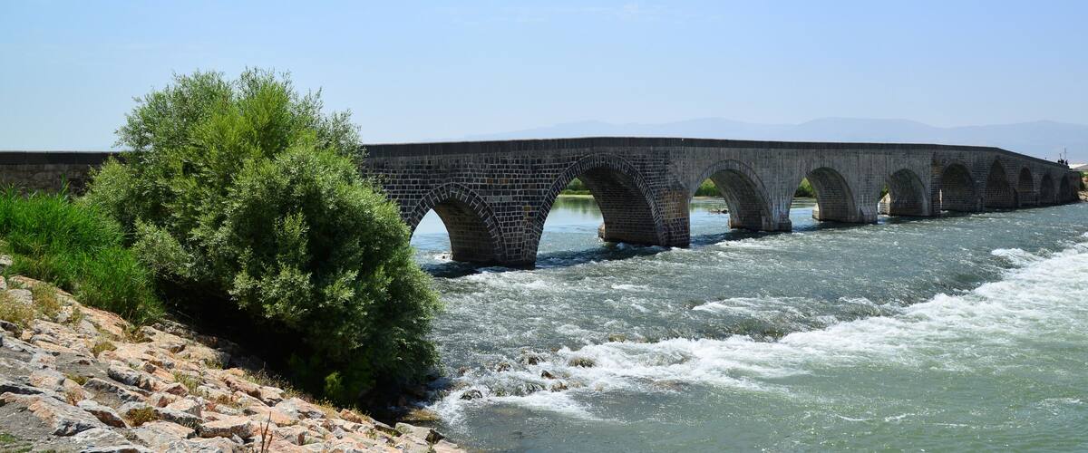 A stone bridge over a river with a rocky riverbank and greenery.