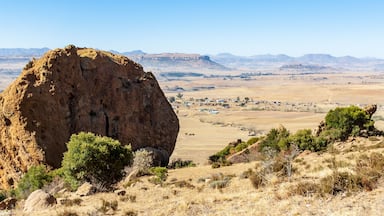 Uma grande formação rochosa em uma paisagem seca e ondulada, com vegetação esparsa e montanhas ao longe.