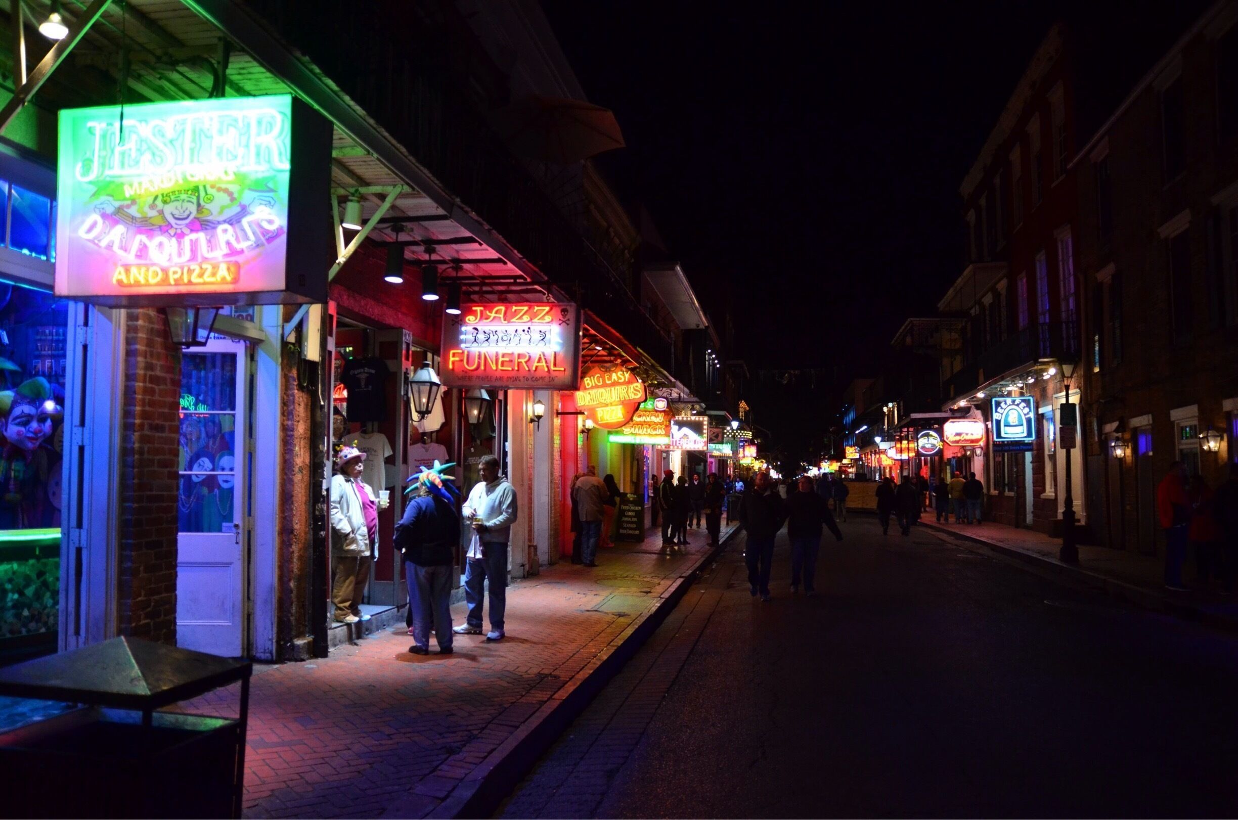 Una calle iluminada de noche con letreros luminosos para una pizzería, un cortejo fúnebre de jazz y otros establecimientos.