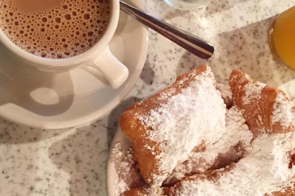 A cup of coffee with a spoon, a glass of water, and a plate of powdered sugar-covered pastries.
