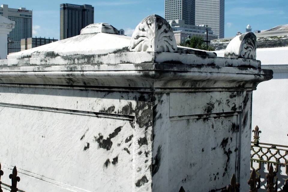 Um monumento desgastado com esculturas de leão, cercado por uma grade de ferro forjado, ao fundo de um horizonte urbano com edifícios modernos.