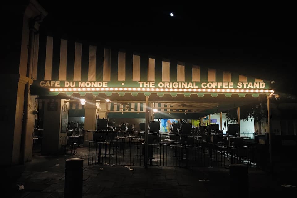A nighttime view of Cafe Du Monde, a historic coffee stand with a striped awning and illuminated sign.