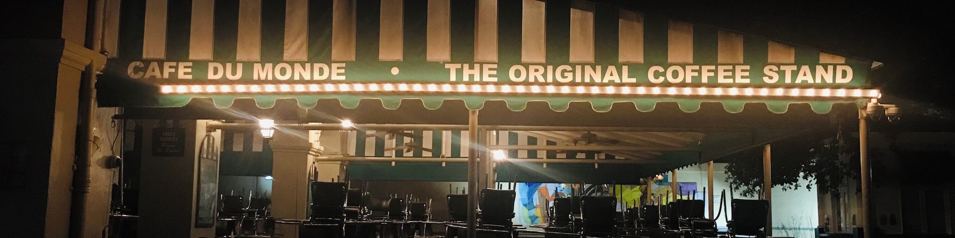 A nighttime view of Cafe Du Monde, a historic coffee stand with a striped awning and illuminated sign.