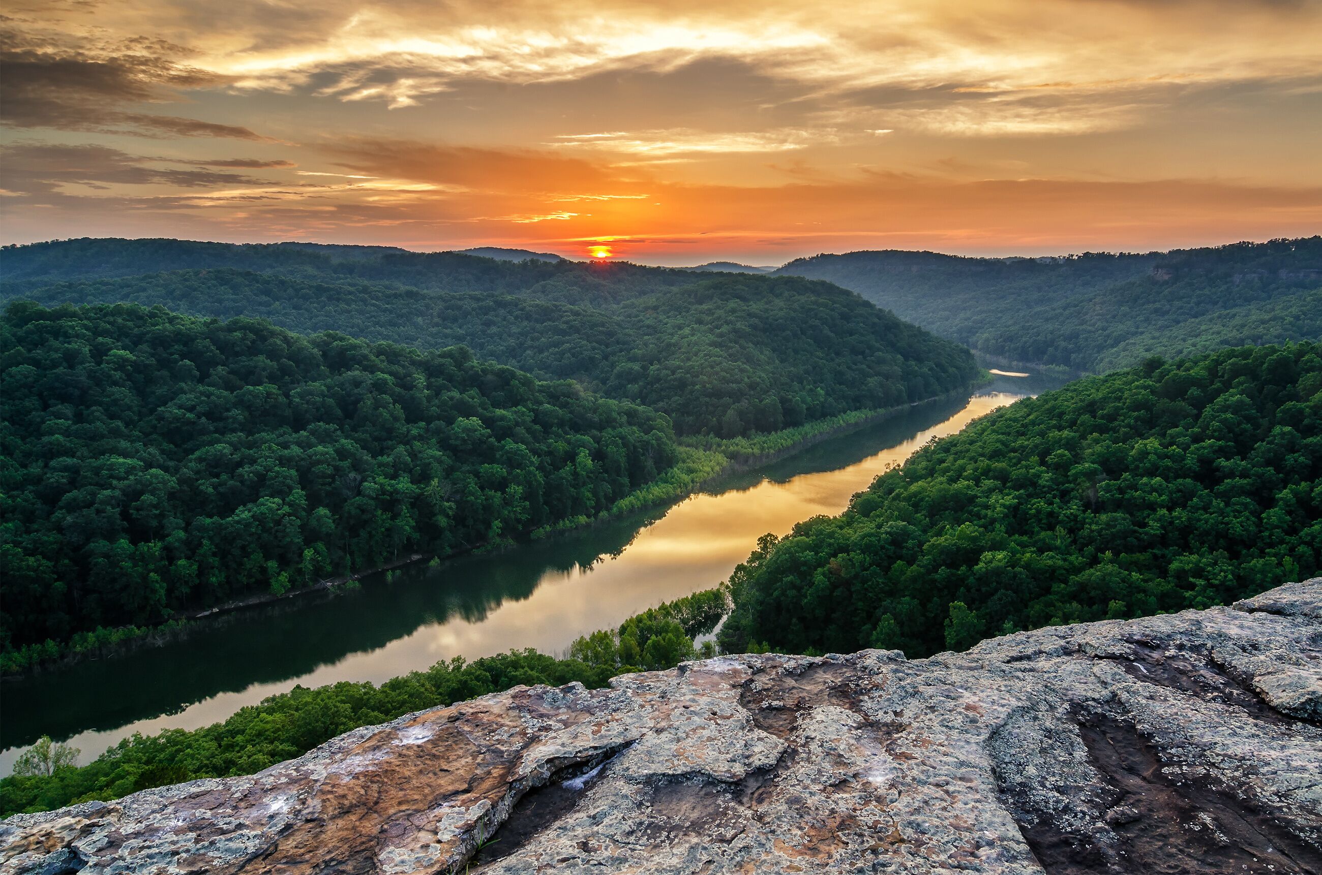 A river winding through a forested valley with a sunset in the background.