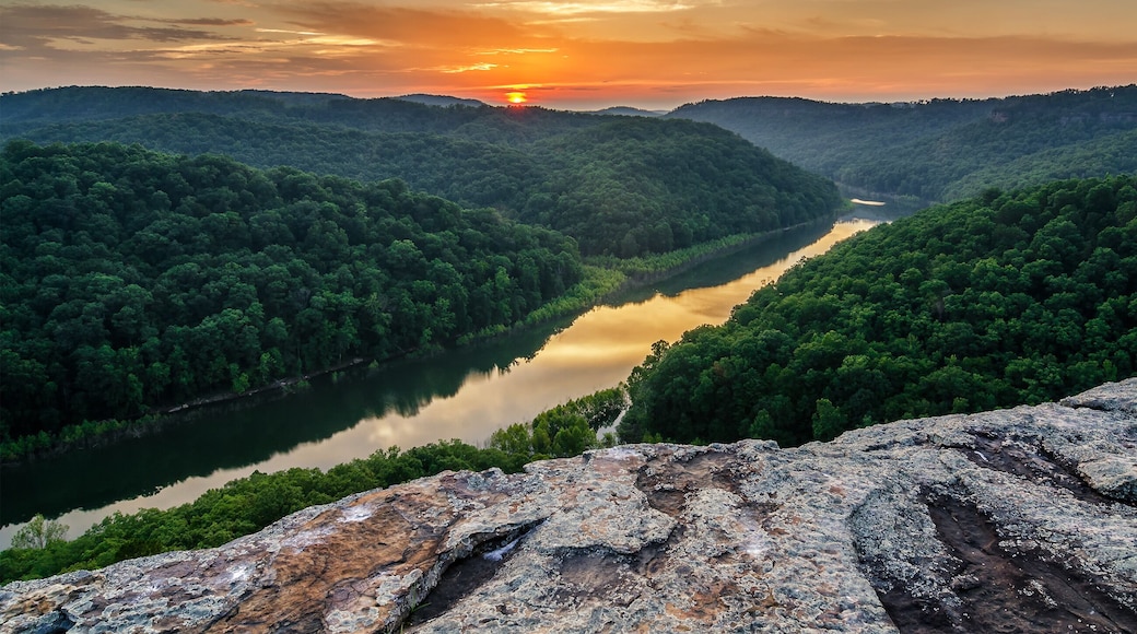A river winding through a forested valley with a sunset in the background.