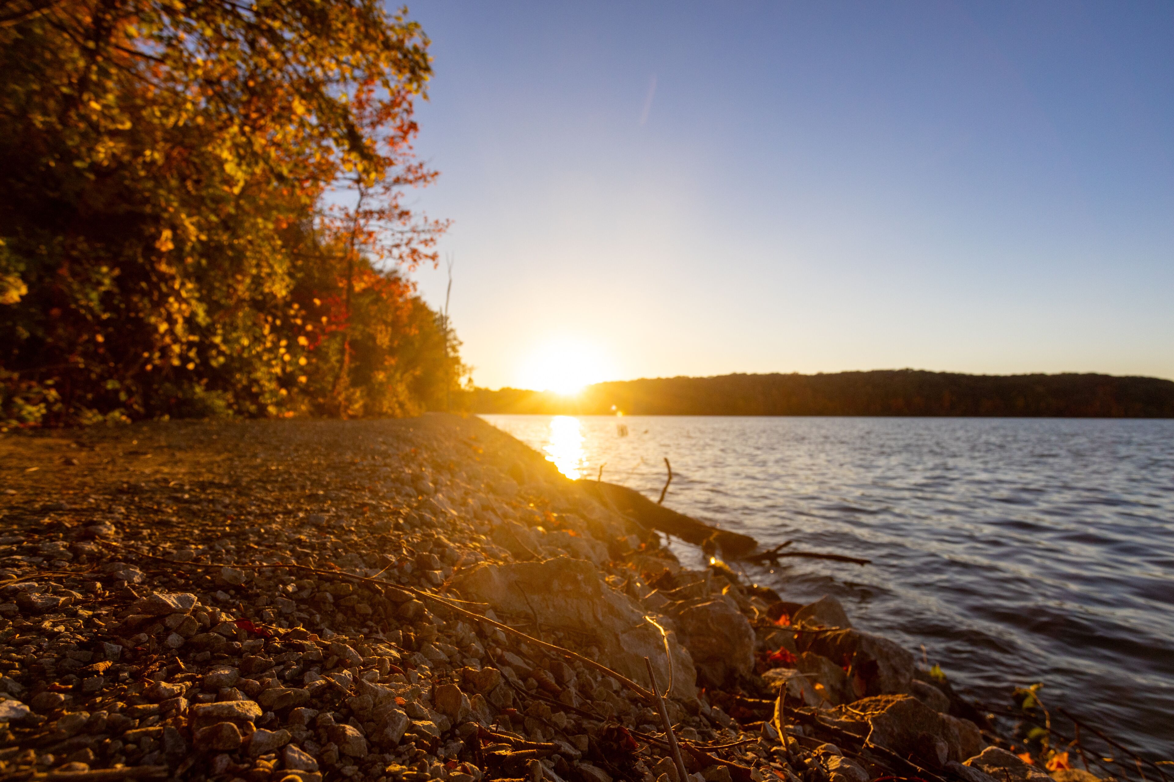 A sunset over a rocky shoreline with trees and a calm lake.