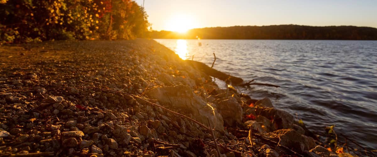 A sunset over a rocky shoreline with trees and a calm lake.