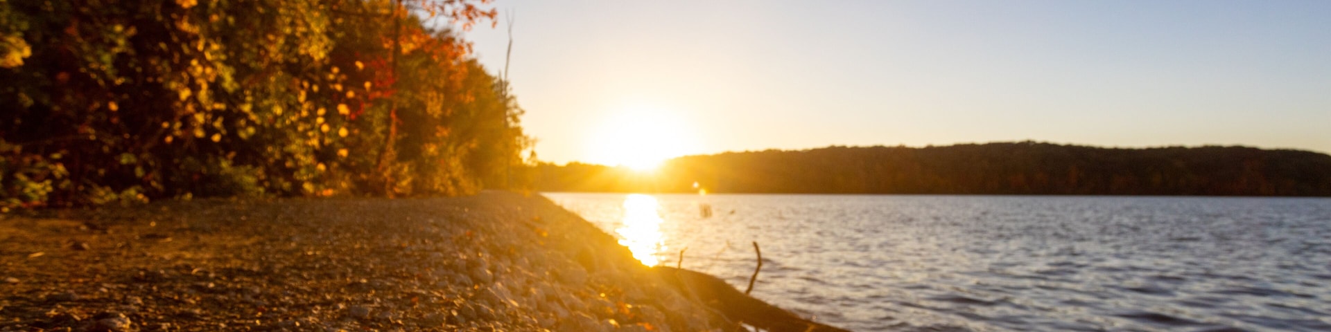 A sunset over a rocky shoreline with trees and a calm lake.