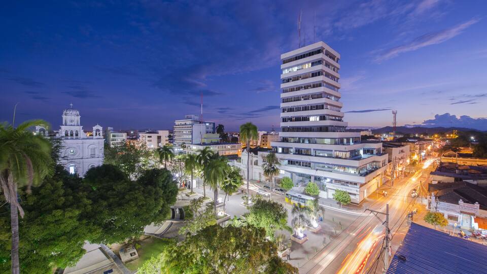 A cityscape at dusk with modern buildings, a historic church, and palm trees.