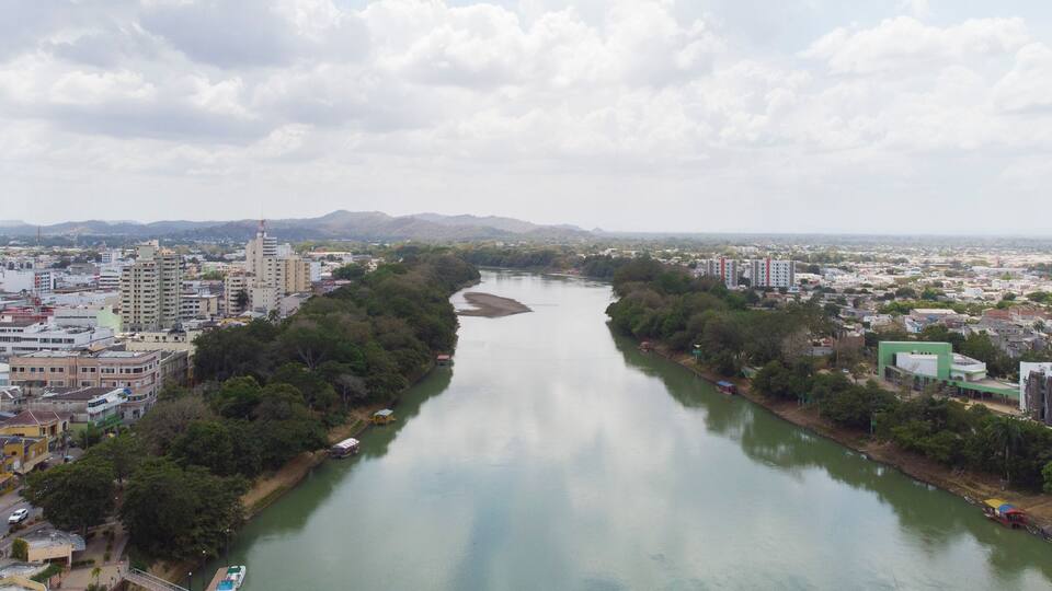 A river flanked by urban buildings and greenery, with mountains in the background.