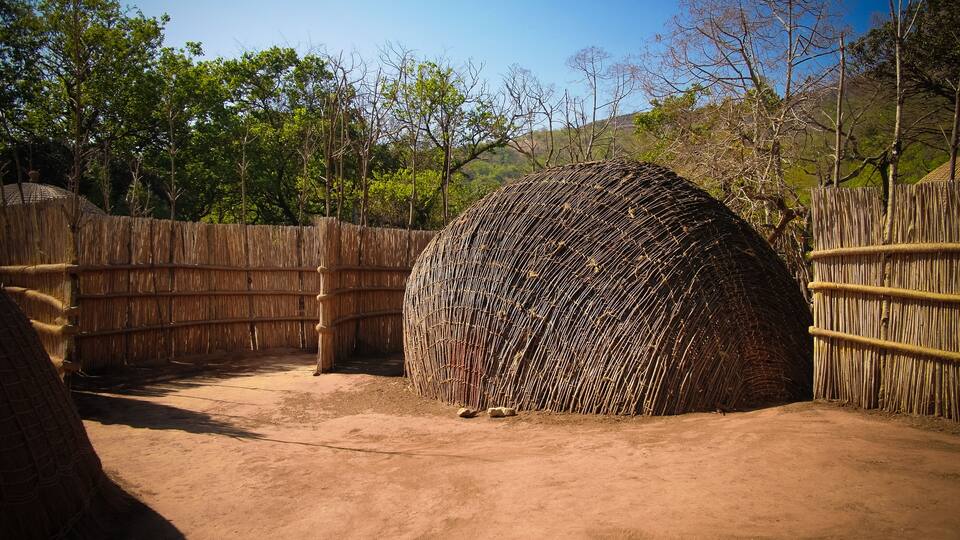 A large, spherical structure made of sticks, surrounded by a bamboo fence in a dry, open area with sparse trees.