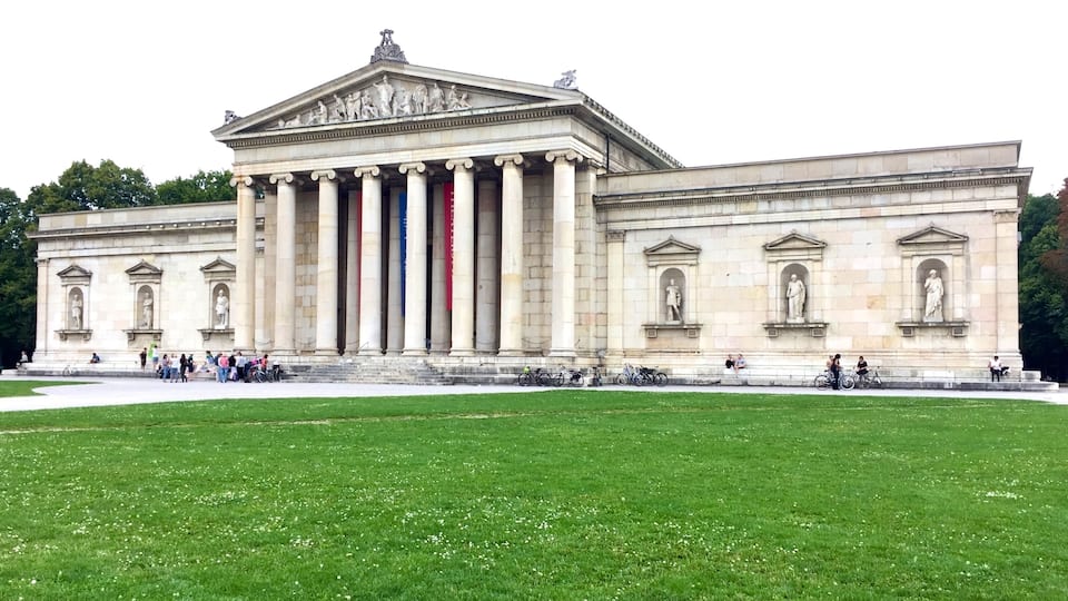 A neoclassical building with large columns and statues, surrounded by a grassy area.