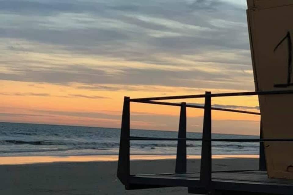 A beach chair on a sandy shore with the ocean and a sunset in the background.