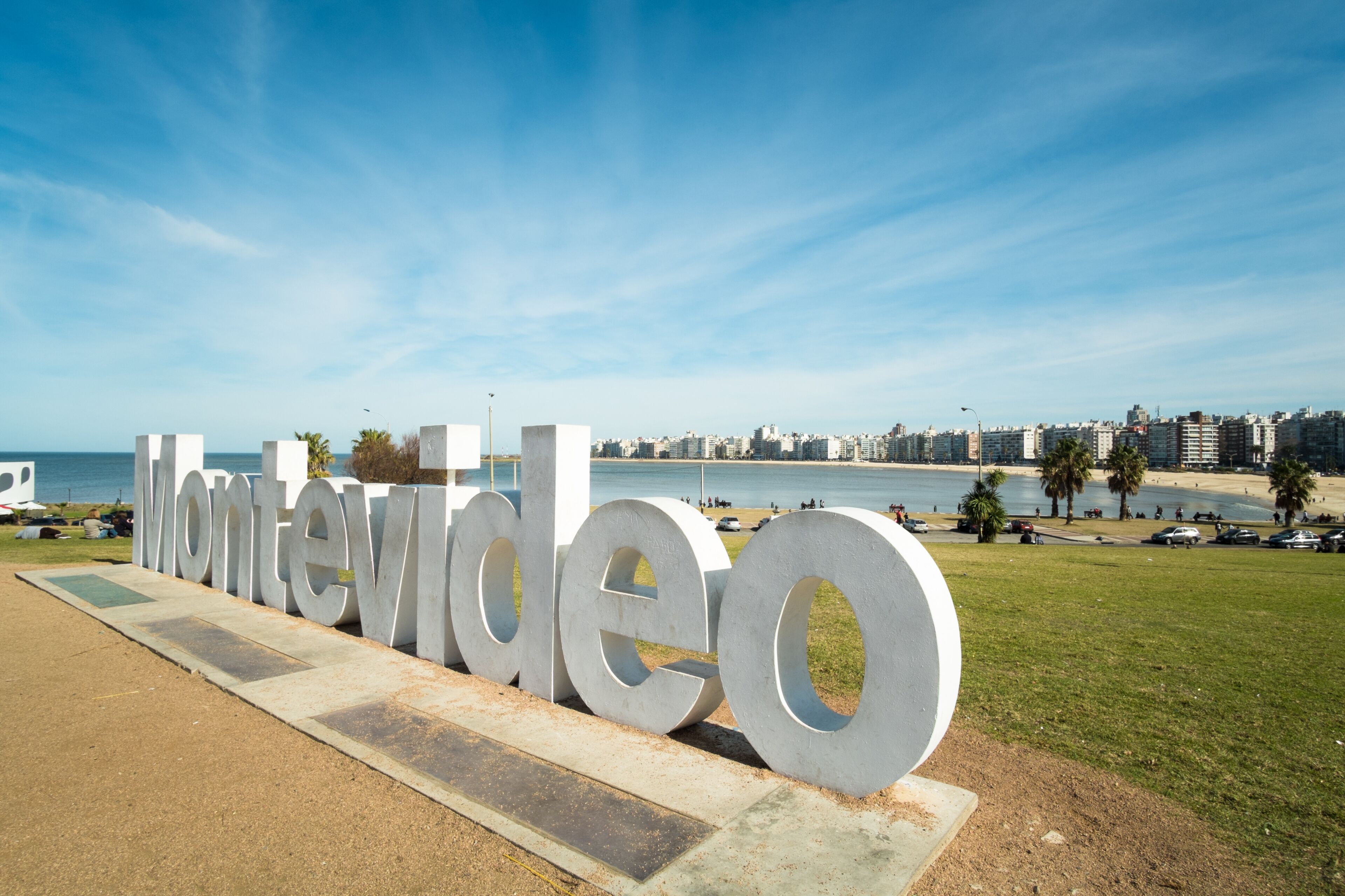 Un letrero grande al aire libre con la palabra 'Montevideo', con un paisaje urbano costero de fondo.