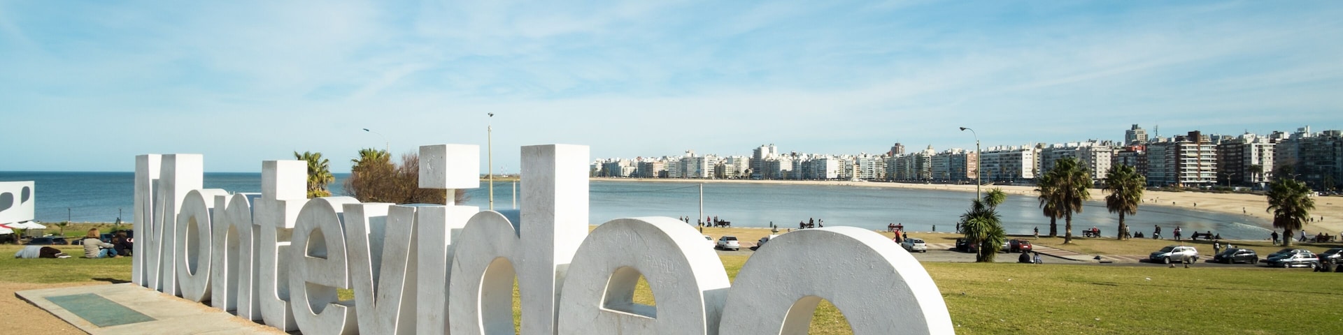 Un letrero grande al aire libre con la palabra 'Montevideo', con un paisaje urbano costero de fondo.